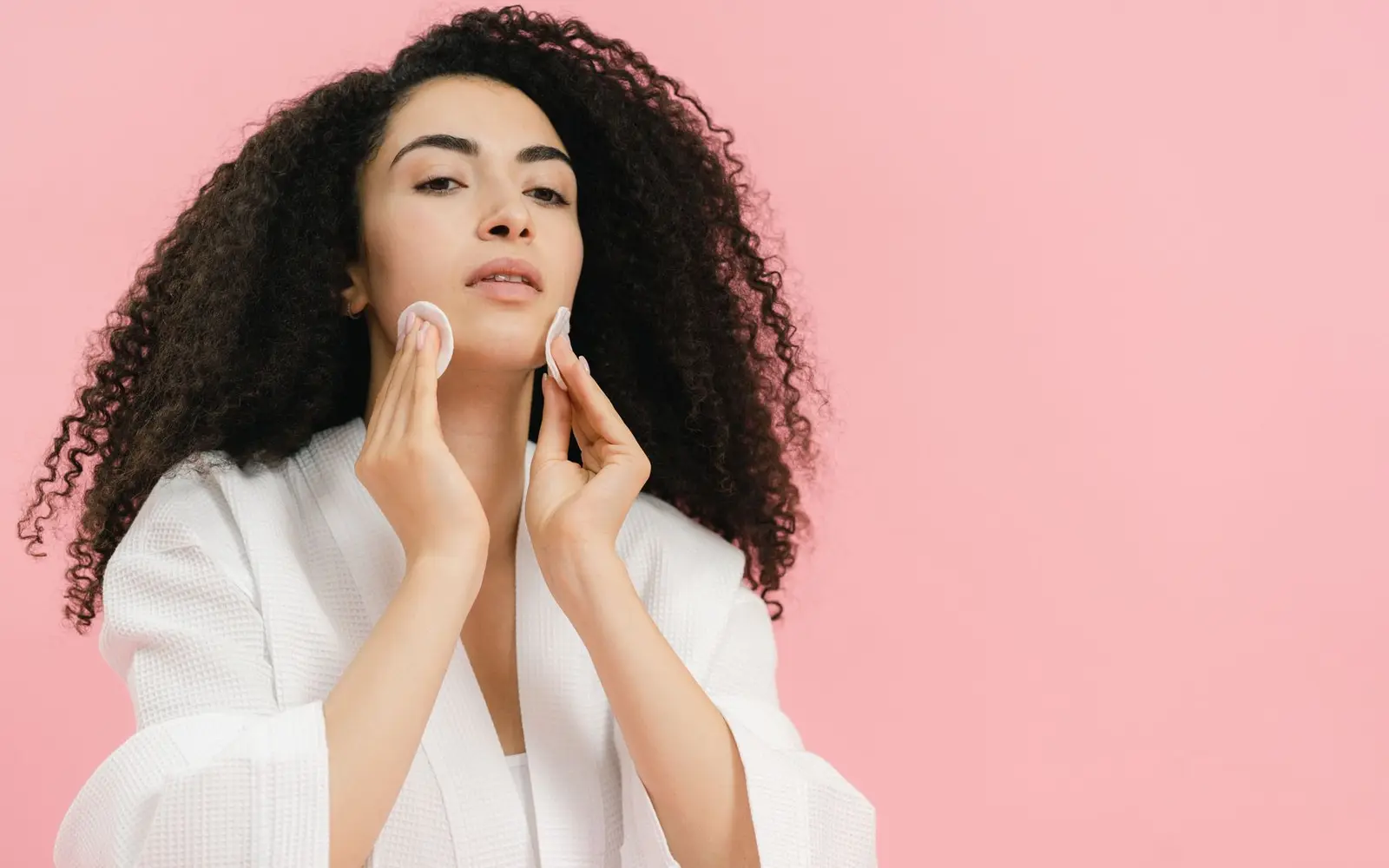 A young woman with curly hair using cotton pads for her skincare routine against a pink background.